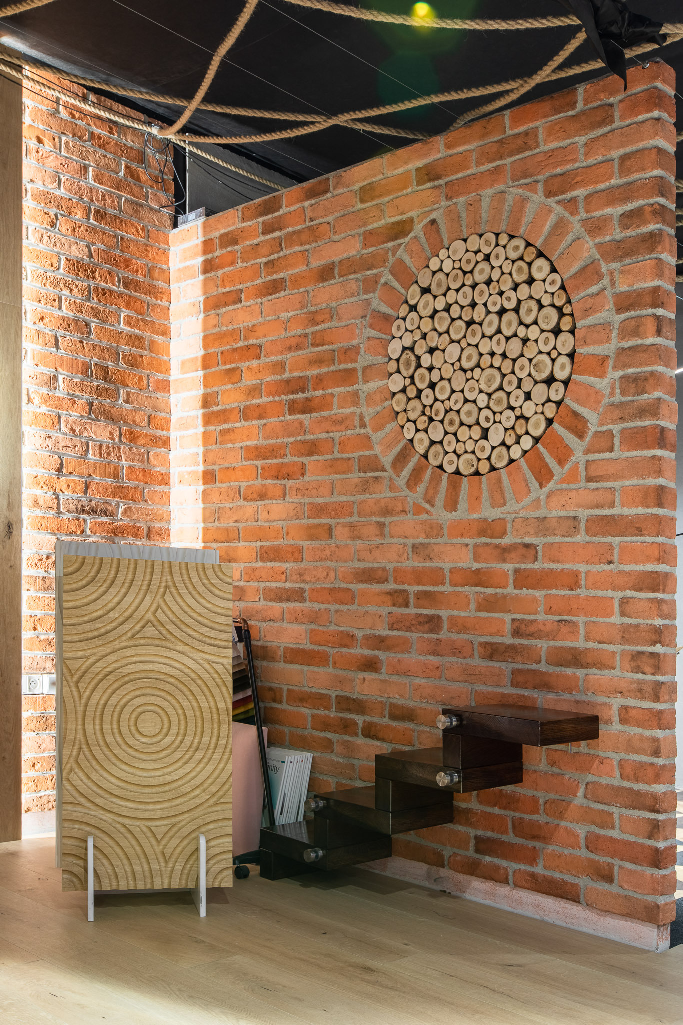 Ceglana ściana z okrągłą dekoracją z drewnianych krążków, obok drewniany panel dekoracyjny z koncentrycznymi okręgami i półkami w formie schodków.