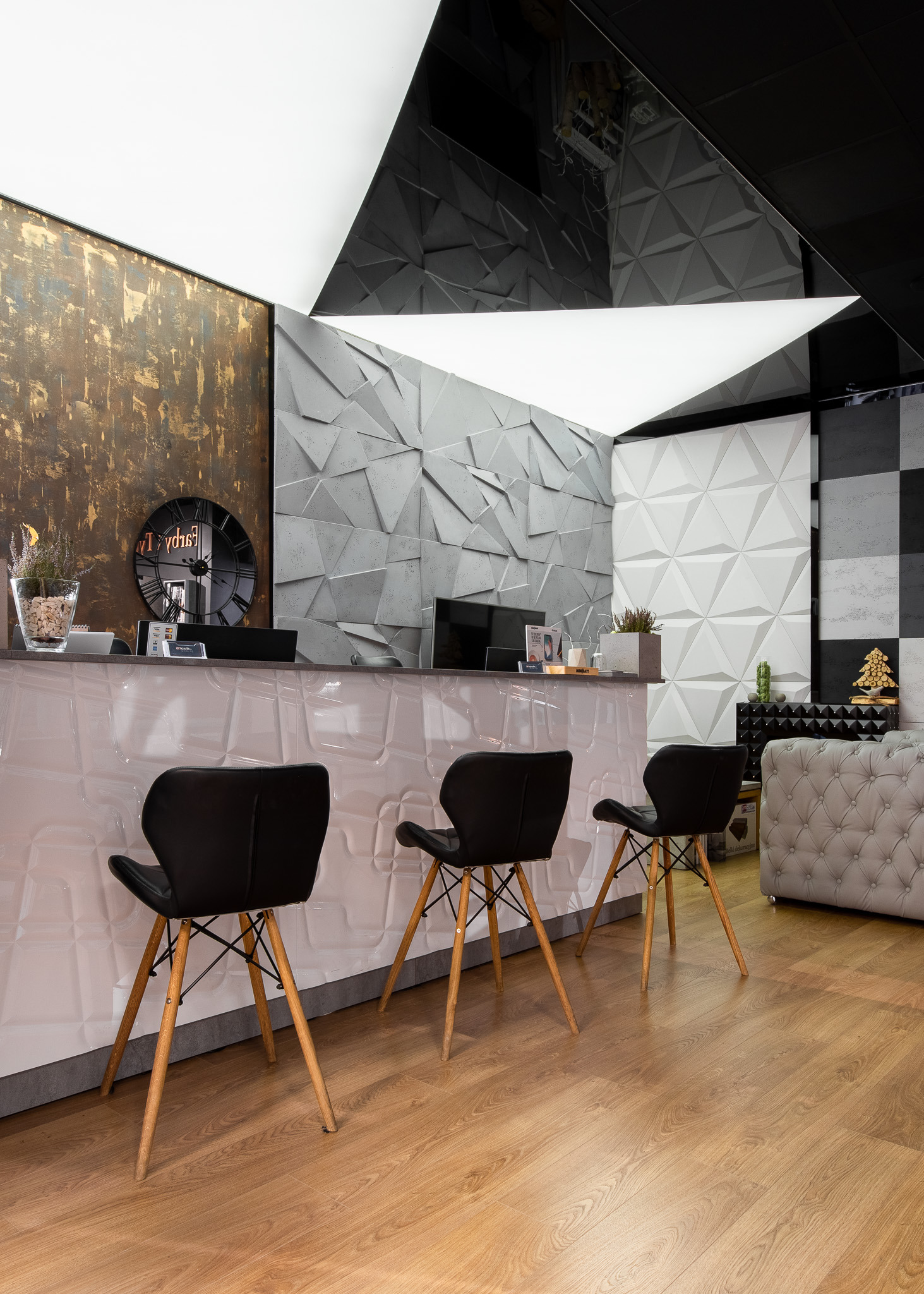Recepcja biurowa z trójkątnymi panelami 3D na ścianie, czarnymi krzesłami na drewnianych nogach i dekoracyjnym zegarem na ścianie w kolorze starego złota.