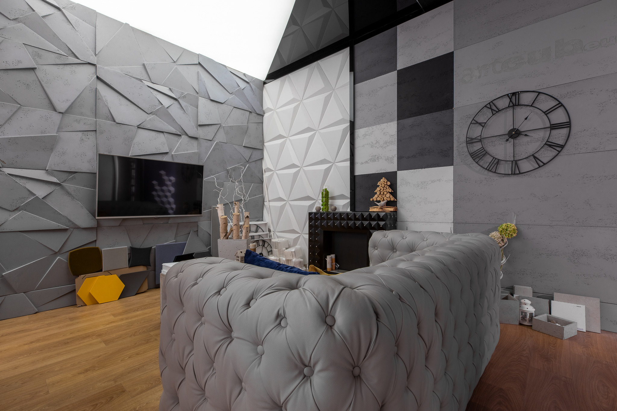 Nowoczesny salon z szarą, pikowaną sofą, trójwymiarową dekoracją ścienną w odcieniach szarości i bieli, oraz dużym, okrągłym zegarem na ścianie.