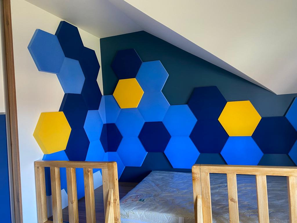 Ściana pokoju dziecięcego z dekoracyjnymi panelami akustycznymi w kształcie sześciokątów w odcieniach niebieskiego i żółtego, widoczne łóżeczko dziecięce z naturalnego drewna.