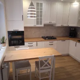 Jasna kuchnia z białymi szafkami i drewnianym blatem, stół z drewnianym blatem i białymi krzesłami, lampa w stylu industrialnym.