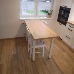 Jasna kuchnia z blatem drewnianym przed oknem, białe szafki z czarnymi uchwytami, drewniana podłoga, minimalistyczny styl.