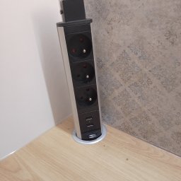 Krystian wojtas Meble Wojtas - Wysuwany z blatu moduł z trzema gniazdkami elektrycznymi i dwoma portami USB, zamontowany w rogu między blatem a ścianą wyłożoną wzorzystą tapetą.