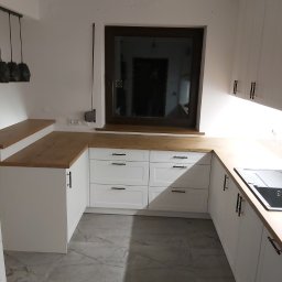 Krystian wojtas Meble Wojtas - Jasna kuchnia z białymi szafkami, drewnianym blatem i szarymi płytkami podłogowymi, widok na okno z ciemną ramą.