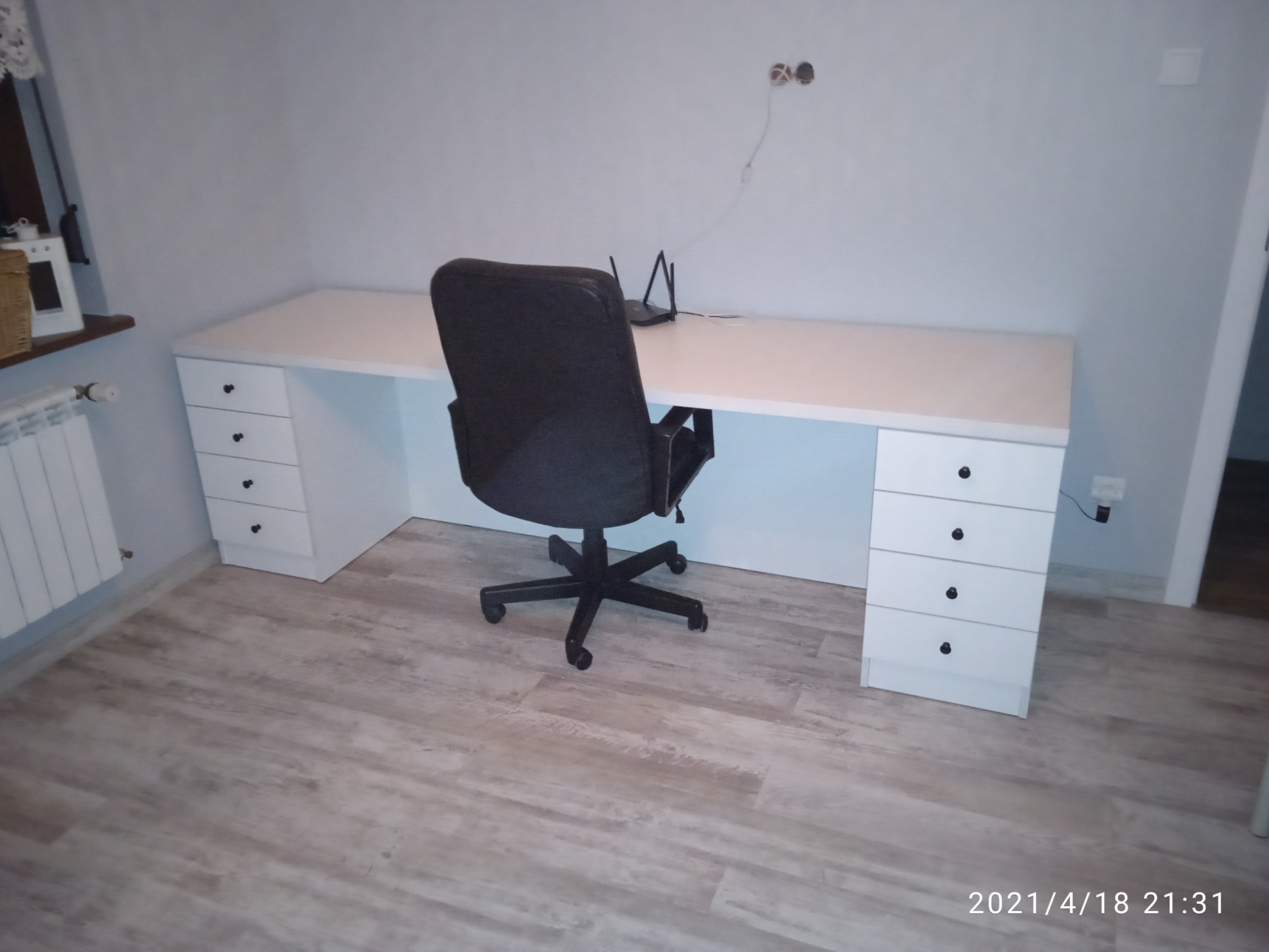 Białe biurko na wymiar z szufladami po bokach, czarne krzesło biurowe, router bezprzewodowy na blacie, drewnopodobna podłoga.