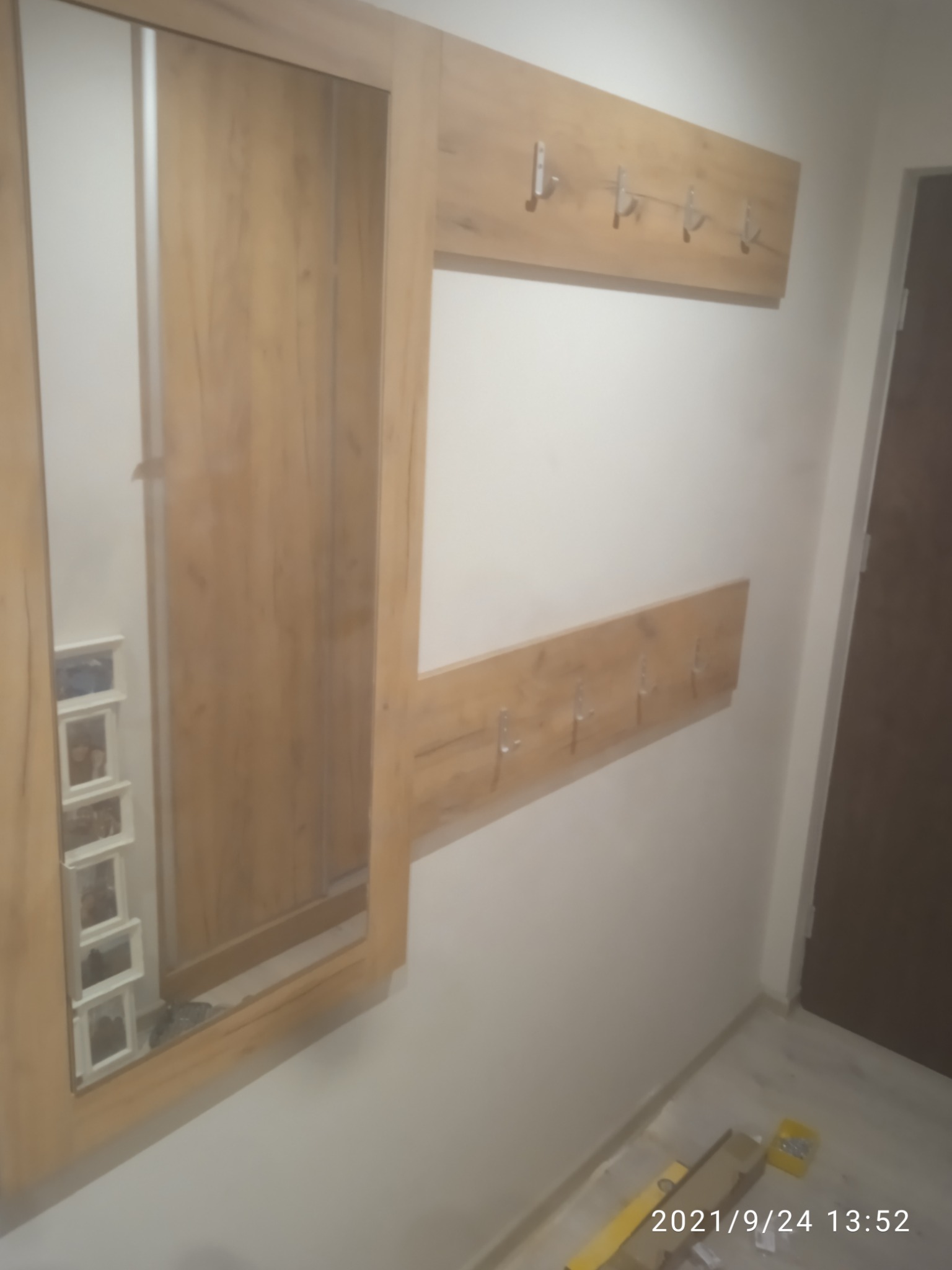 Fragment przedpokoju z szafą z lustrem i dwoma panelami z wieszakami w kolorze drewna, widoczne narzędzia na podłodze.