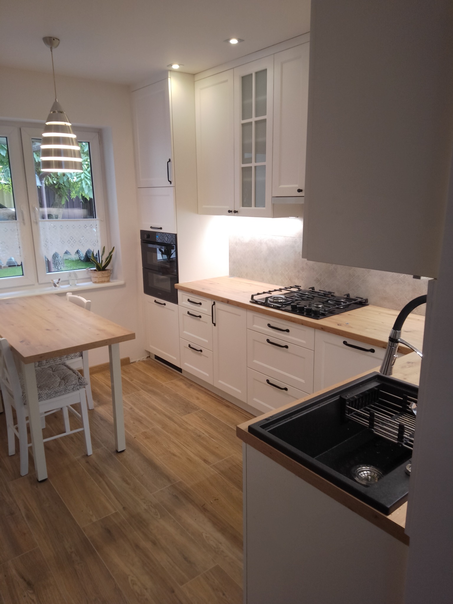 Jasna kuchnia na wymiar z drewnianym blatem, czarnym zlewem i płytą gazową, widok na okno z roletą i stół z krzesłami.