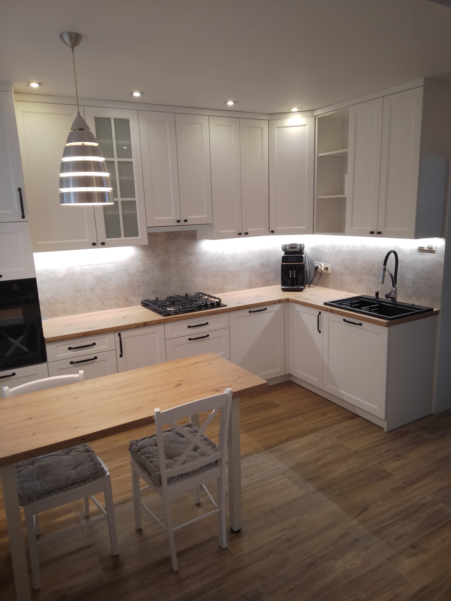 Jasna kuchnia z białymi szafkami, drewnianymi blatami i stołem z krzesłami w tle, oświetlona lampą wiszącą i światłem LED pod szafkami.