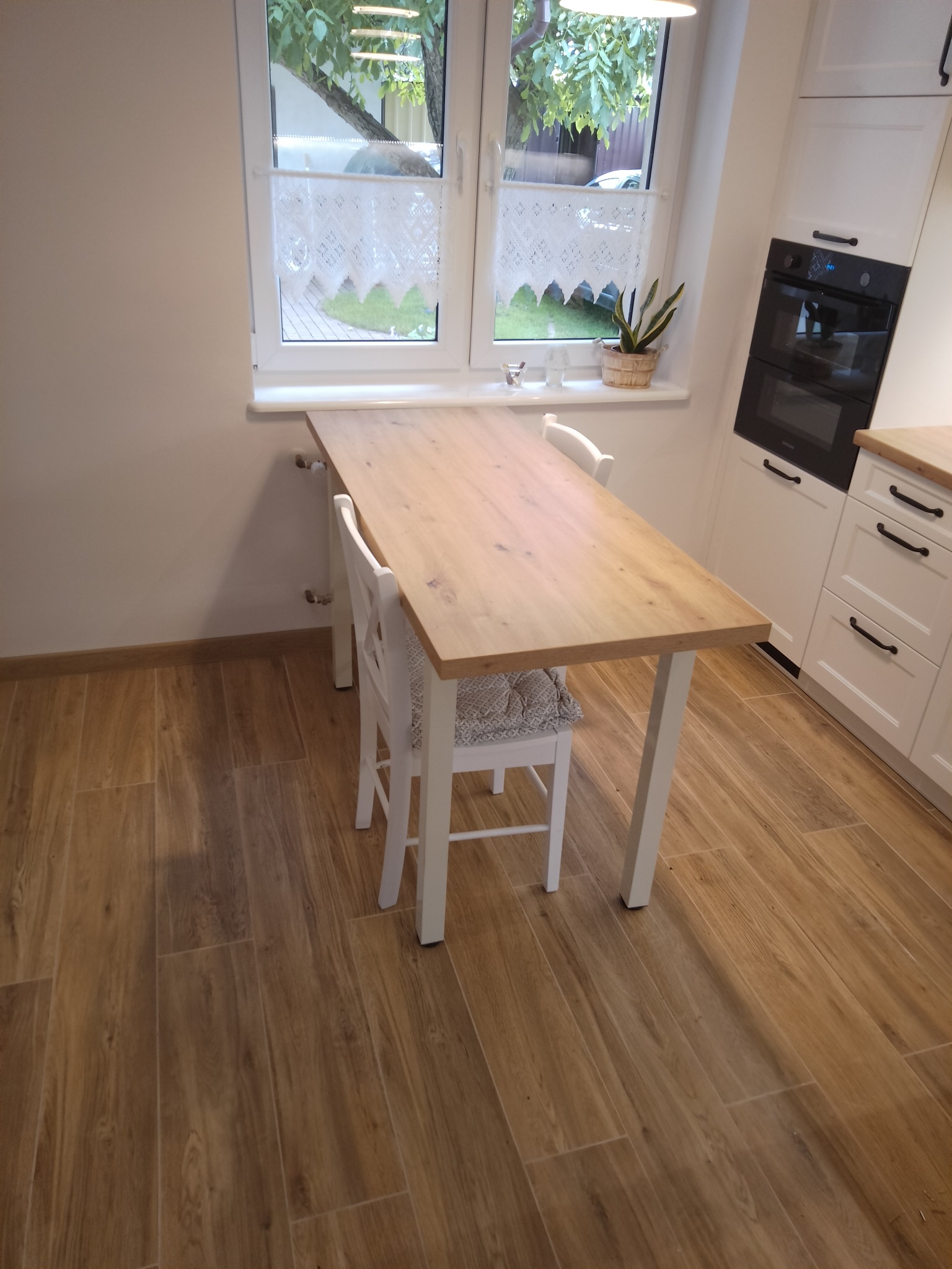 Jasna kuchnia z blatem drewnianym przed oknem, białe szafki z czarnymi uchwytami, drewniana podłoga, minimalistyczny styl.