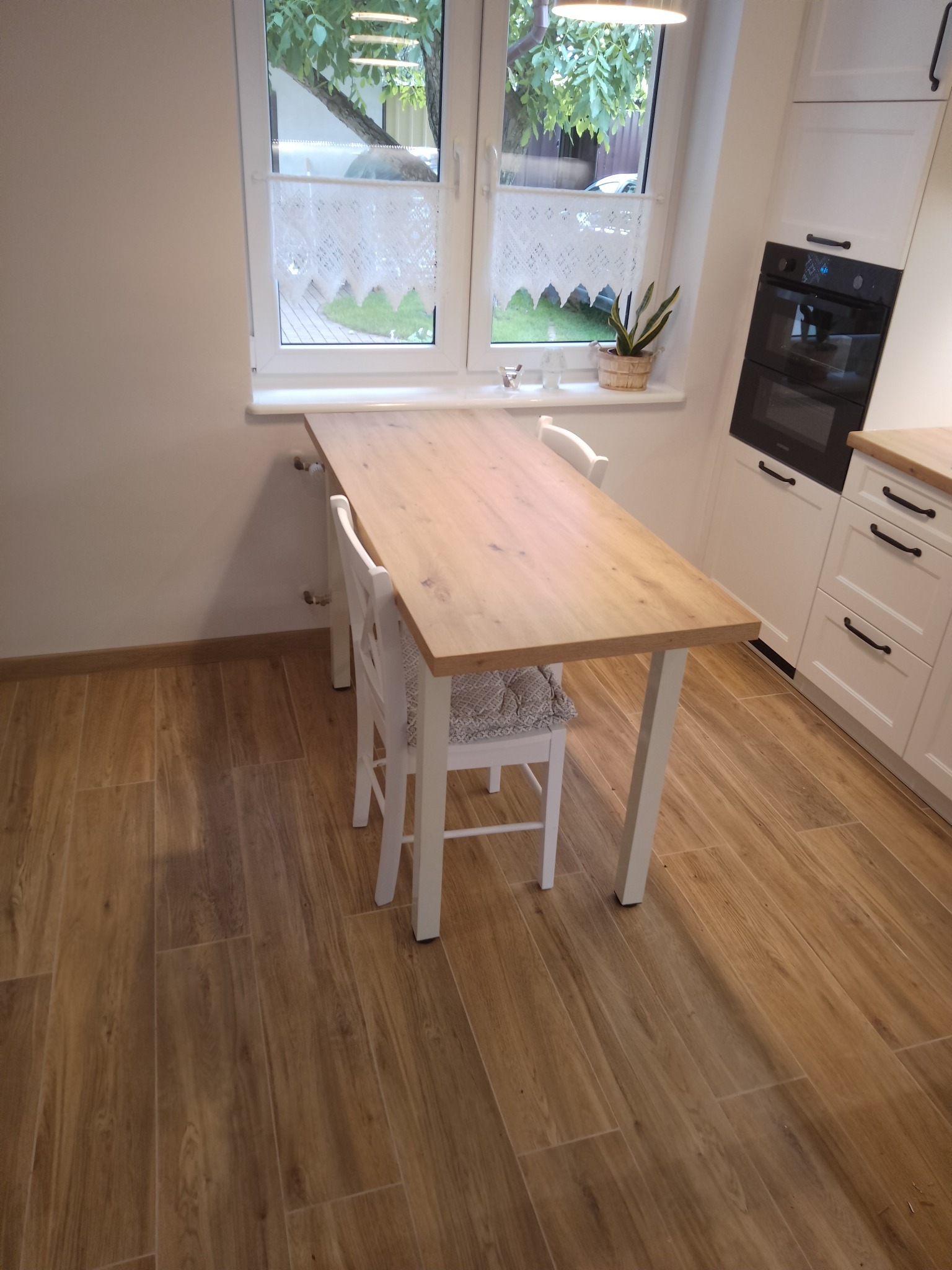 Jasna kuchnia z drewnianym blatem stołu, białymi krzesłami i szafkami, widok na okno z koronkową firanką i rośliną doniczkową.