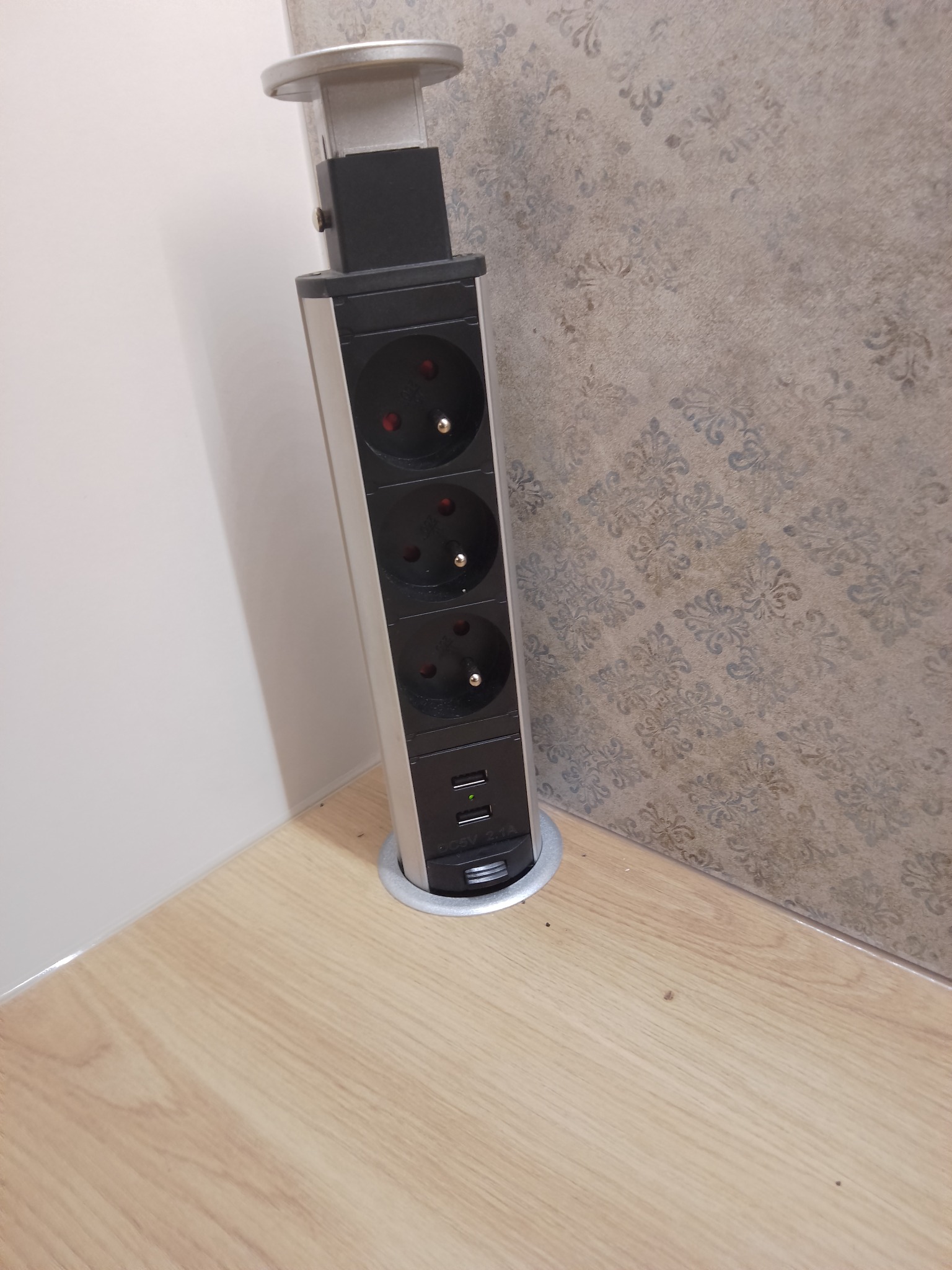 Wysuwany z blatu moduł z trzema gniazdkami elektrycznymi i dwoma portami USB, zamontowany w rogu między blatem a ścianą wyłożoną wzorzystą tapetą.