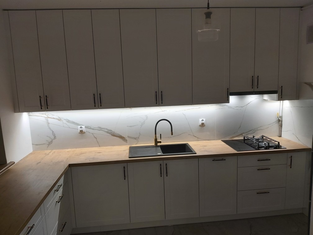 Biała kuchnia na wymiar z drewnianym blatem, czarnym zlewem i płytą gazową, podświetlana listwą LED pod szafkami wiszącymi, z marmurowym wzorem na ścianie.