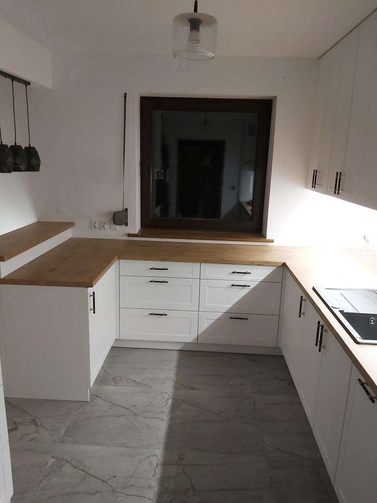 Jasna kuchnia z białymi szafkami, drewnianym blatem i szarymi płytkami podłogowymi, widok na okno z ciemną ramą.