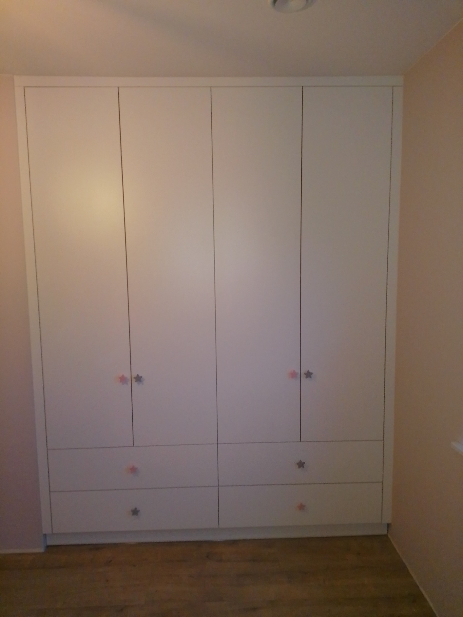 Biała szafa wnękowa z czterema drzwiami i czterema szufladami, uchwyty w kształcie gwiazdek w dwóch kolorach, różowym i szarym, na tle ściany w odcieniu pudrowego różu.