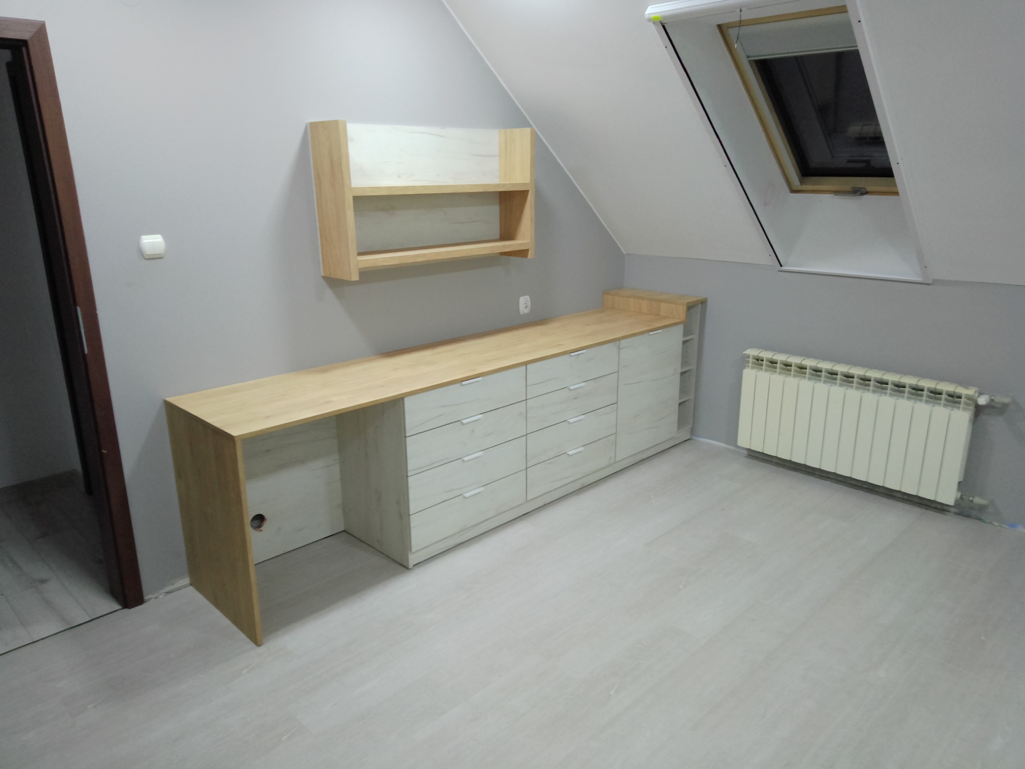 Szafki i blat z jasnego drewna na poddaszu ze skosem, szuflady z białymi uchwytami, półka wisząca, grzejnik pod oknem dachowym.