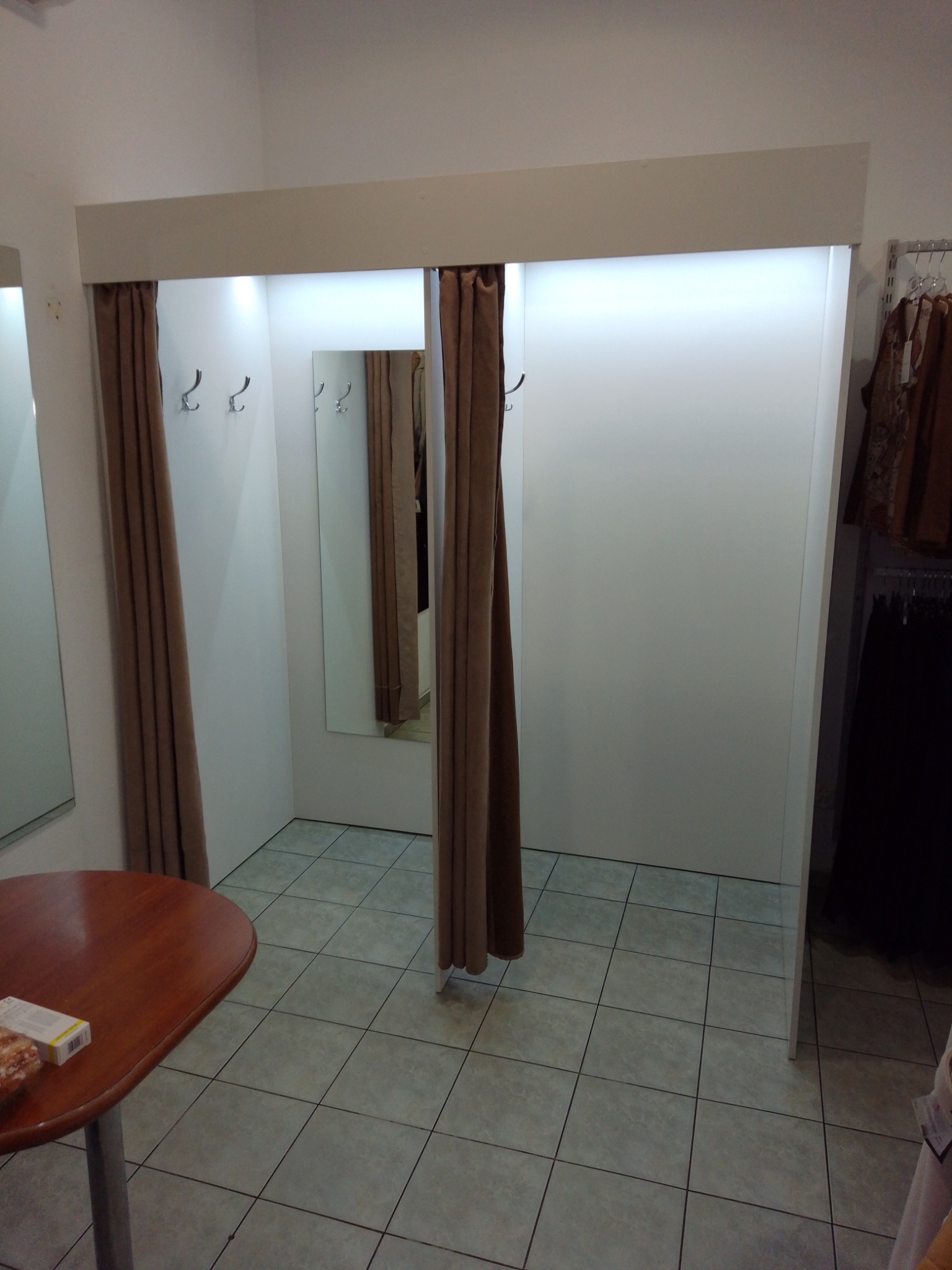 Trzy kabiny przymierzalni z brązowymi zasłonami, lustrami i wieszakami, widoczne fragmenty ubrań na wieszakach obok.