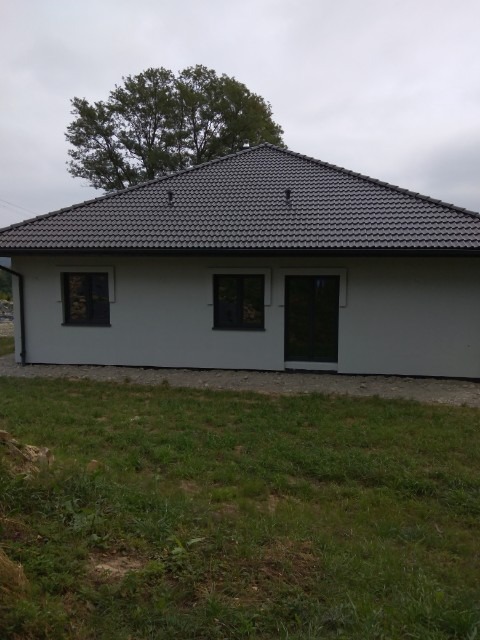 Dom jednorodzinny z nowym, ciemnoszarym dachem z dachówki ceramicznej, widoczny z poziomu trawnika w pochmurny dzień.