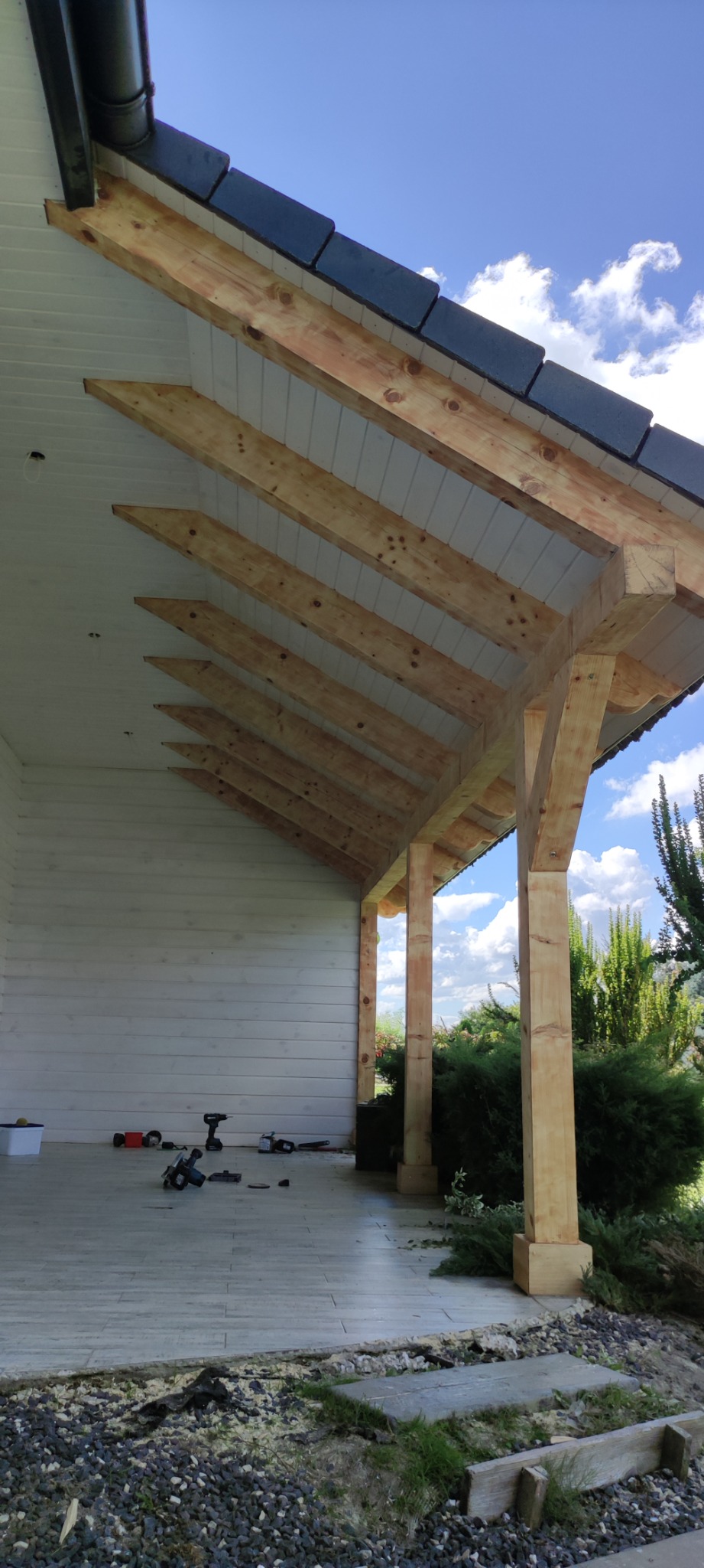 Konstrukcja drewniana z widoczną więźbą dachową, częściowo pokrytą ciemną dachówką, wsparta na drewnianych słupach, z narzędziami budowlanymi leżącymi na podłodze.