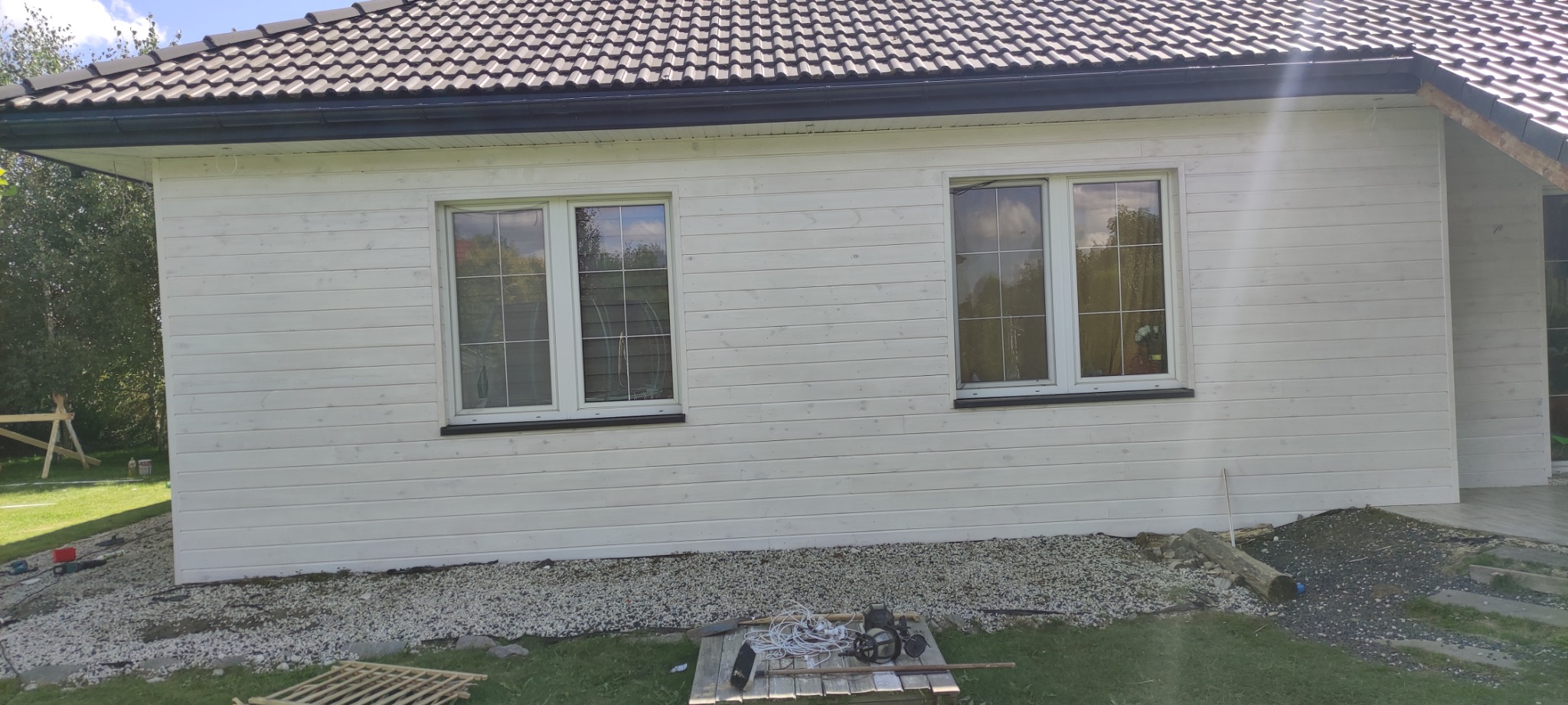 Ściana domu z drewnianą elewacją pomalowaną na biało, z dwoma oknami i ciemnym dachem z rynnami. Na trawie widoczne narzędzia i materiały budowlane.