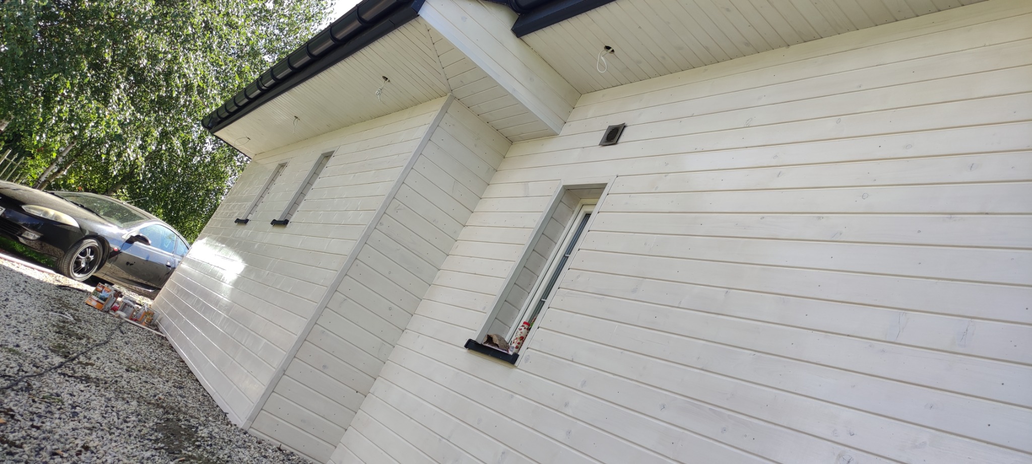 Świeżo pomalowana na biało drewniana elewacja domu z widoczną czarną rynną, samochodem i puszkami farby na żwirowym podłożu.