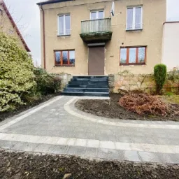 Szara ścieżka z kostki brukowej z jasnymi obrzeżami prowadząca do schodów wejściowych domu z beżową elewacją i brązowymi oknami, widok na krzewy i drzewa w tle.