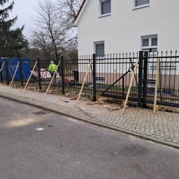 Ivan Kletsyk - Montaż czarnego ogrodzenia metalowego z bramą na posesji, widoczne podpory z drewna, pracownik w odblaskowej kamizelce i narzędzia.