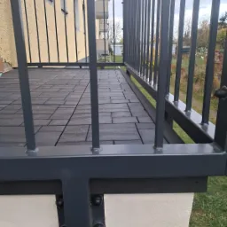 Szare metalowe balustrady balkonu z podłogą wyłożoną ciemnoszarymi płytkami, widok z poziomu podłogi na budynek w tle.