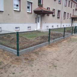 Ivan Kletsyk - Zielona siatka ogrodzeniowa panelowa na betonowej podmurówce, widok z zewnątrz posesji, ziemia wokół ogrodzenia częściowo przekopana, w tle budynek mieszkalny z czerwonymi akcentami.