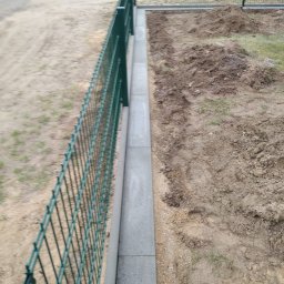 Ivan Kletsyk - Zielona siatka ogrodzeniowa zamontowana na betonowej podmurówce, widok z boku na niezagospodarowany teren.