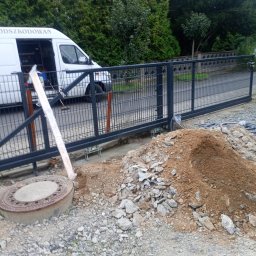 Ivan Kletsyk - Montaż przesuwnej bramy z paneli metalowych w trakcie prac ziemnych, widoczny samochód dostawczy i sterta ziemi.
