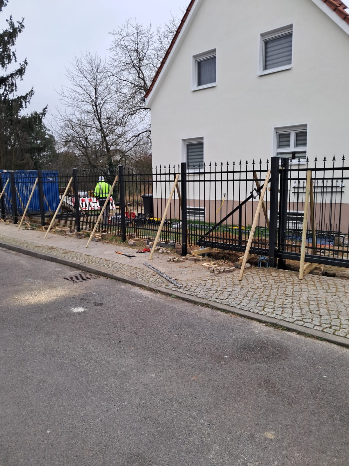 Montaż czarnego ogrodzenia metalowego z bramą na posesji, widoczne podpory z drewna, pracownik w odblaskowej kamizelce i narzędzia.