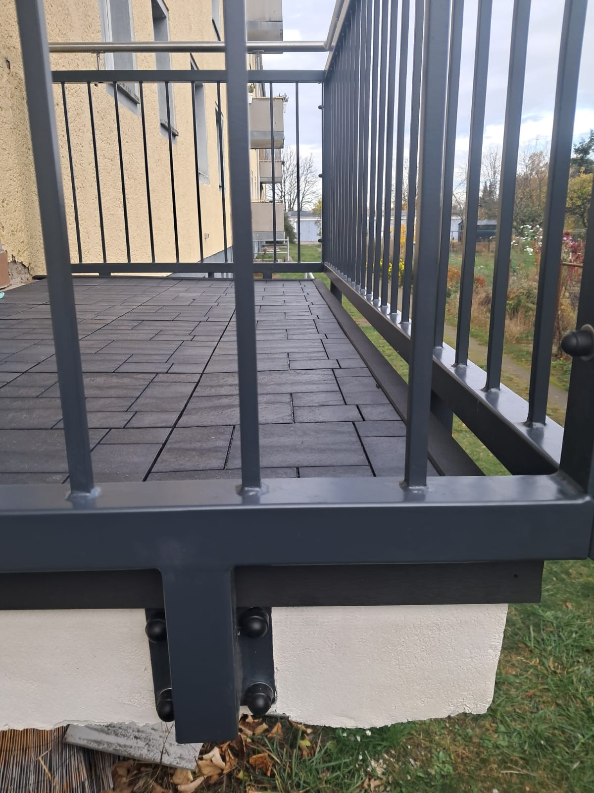 Szare metalowe balustrady balkonu z podłogą wyłożoną ciemnoszarymi płytkami, widok z poziomu podłogi na budynek w tle.