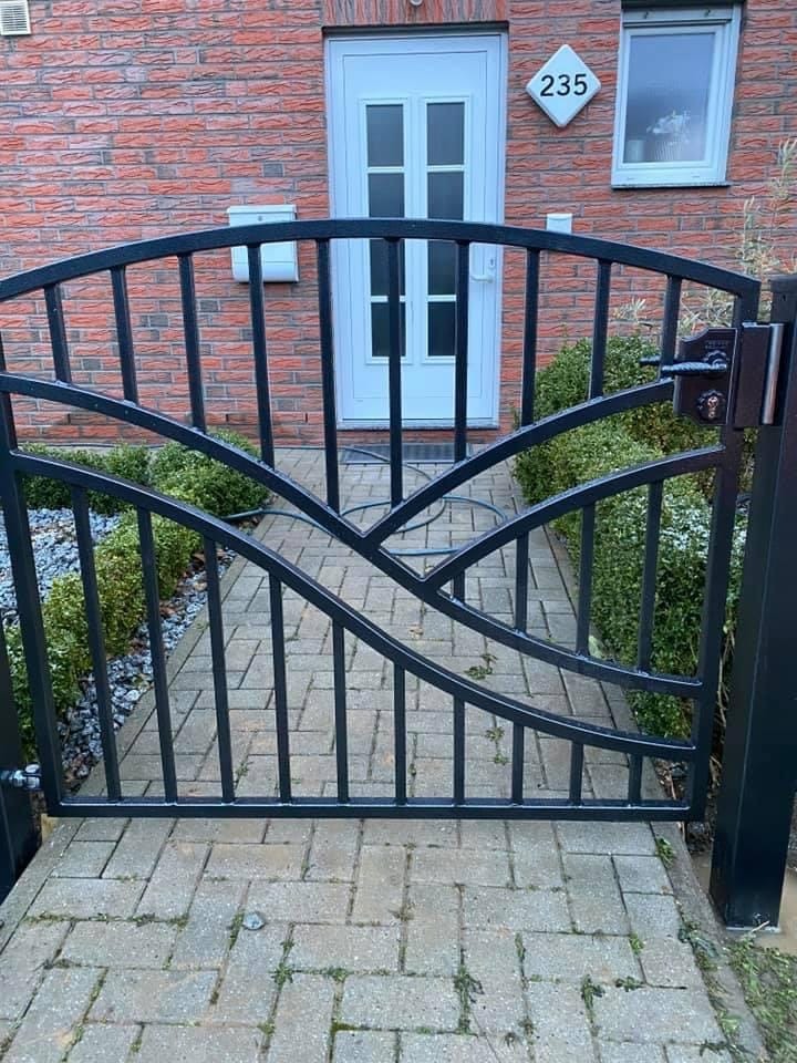 Czarna, metalowa furtka z dekoracyjnym, łukowatym wzorem, prowadząca do domu z ceglaną elewacją i białymi drzwiami.
