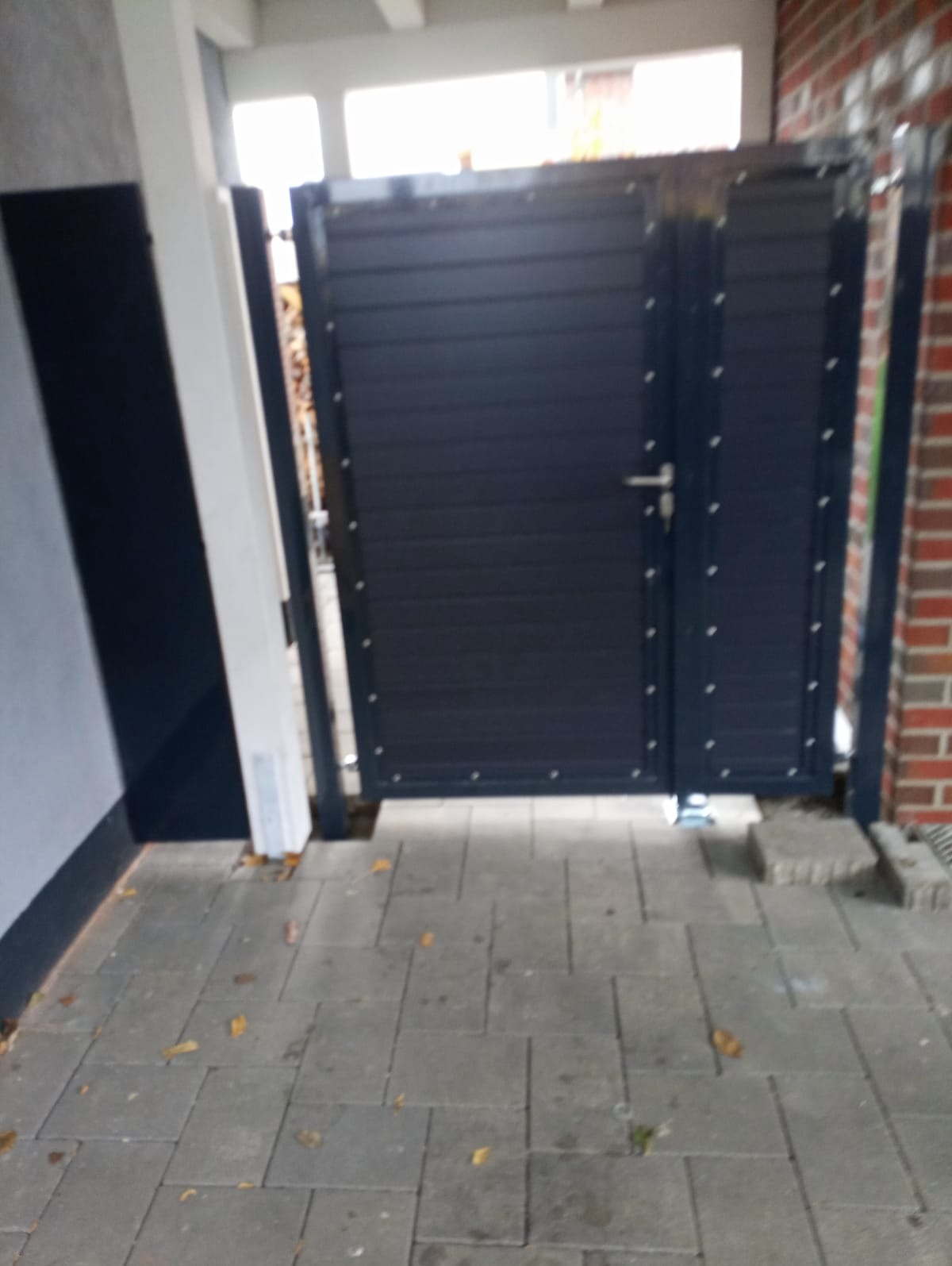 Zamknięta, nowoczesna, dwuskrzydłowa brama metalowa w kolorze ciemnografitowym, z poziomymi panelami, zamontowana przy posesji z kostki brukowej i ceglanego muru.