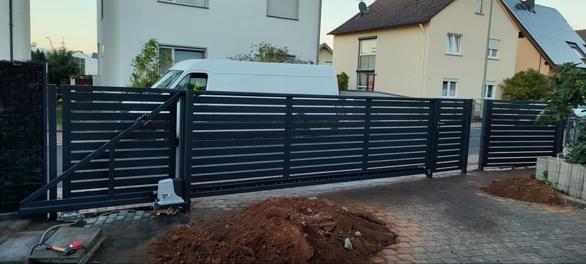 Nowoczesna, przesuwna brama z poziomych, metalowych profili, z automatyką, widoczna sterta ziemi po montażu, na tle dwóch domów i białego samochodu dostawczego.