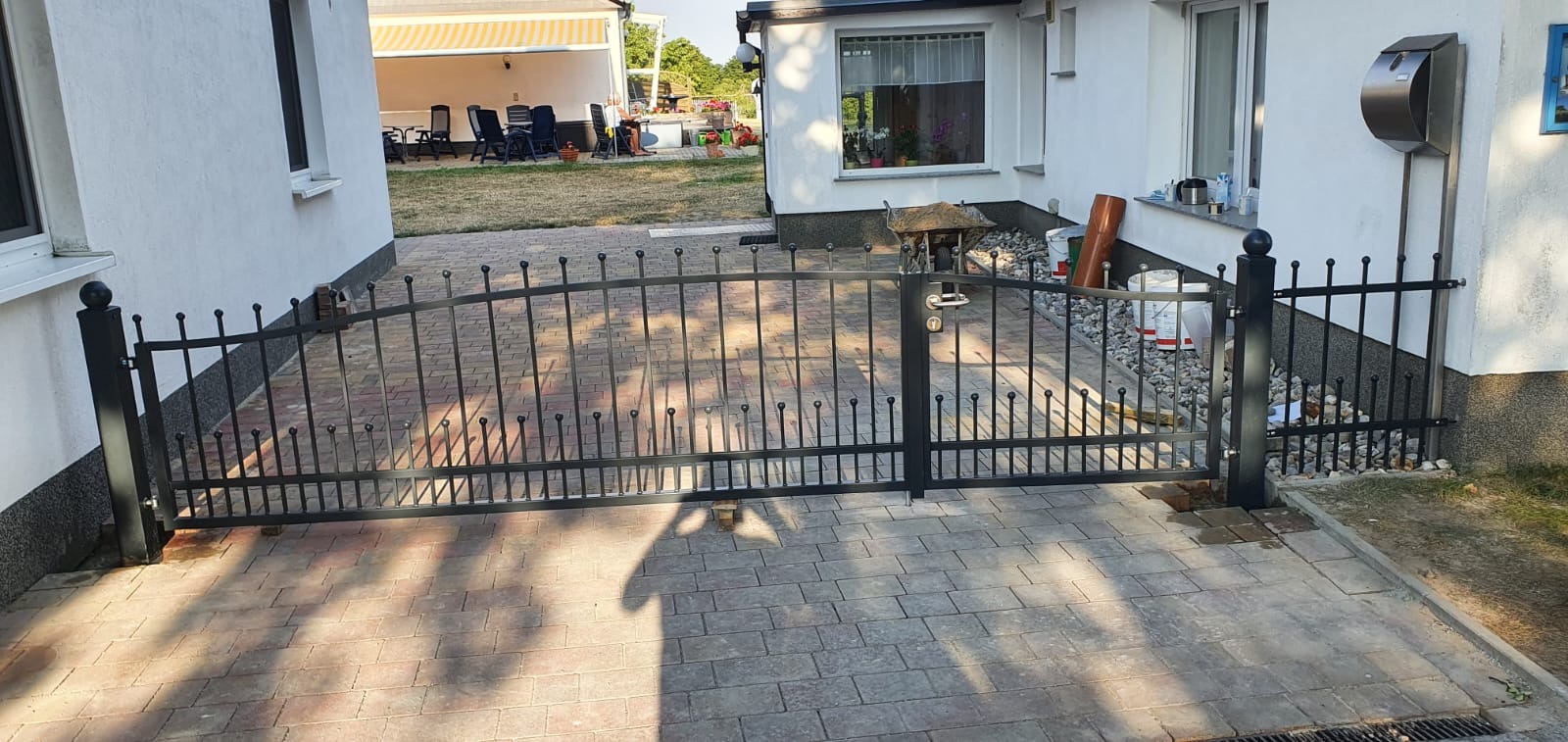 Czarna, metalowa brama z zaokrąglonym łukiem na wjeździe do posesji, widoczna z perspektywy podjazdu wyłożonego kostką brukową, z domem w tle.