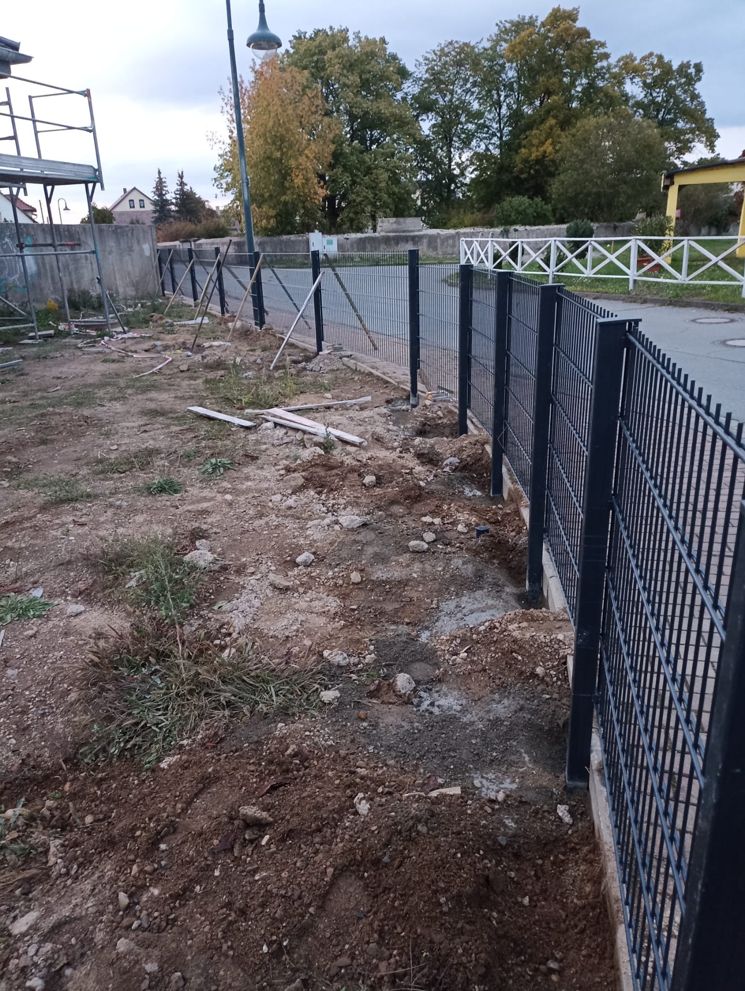 Fragment montażu ciemnego ogrodzenia panelowego na nierównym terenie, widoczne słupki i panele, w tle budowa i drzewa.