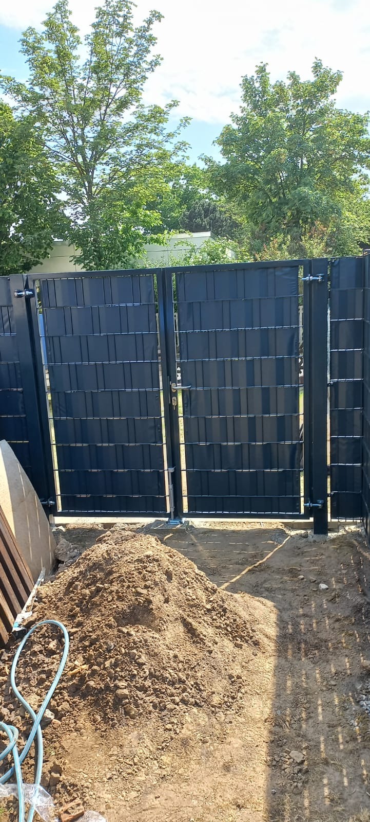 Brama w trakcie montażu z widoczną stertą ziemi i niebieskim wężem ogrodowym, na tle zieleni drzew.