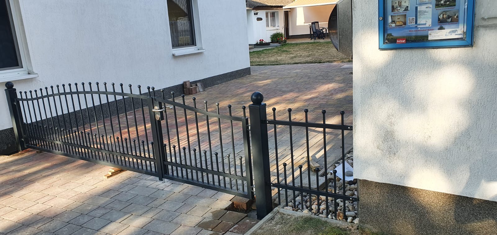 Czarna, metalowa brama z dekoracyjnymi elementami na posesji z kostki brukowej, widok z boku, w tle białe budynki.