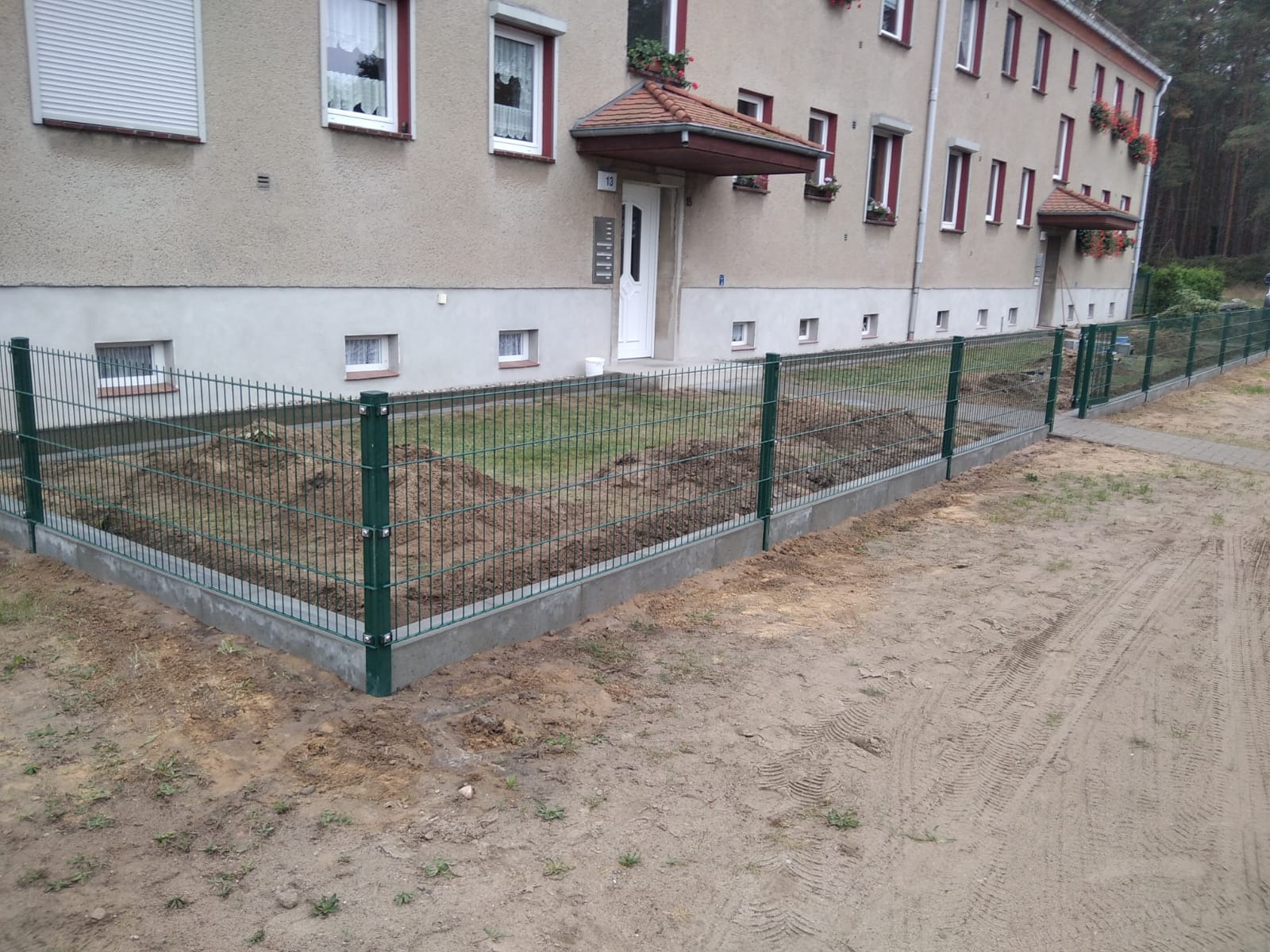 Zielona siatka ogrodzeniowa panelowa na betonowej podmurówce, widok z zewnątrz posesji, ziemia wokół ogrodzenia częściowo przekopana, w tle budynek mieszkalny z czerwonymi akcentami.