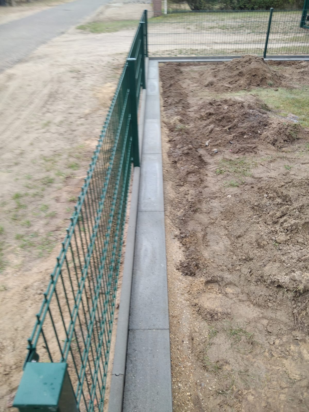 Zielona siatka ogrodzeniowa zamontowana na betonowej podmurówce, widok z boku na niezagospodarowany teren.