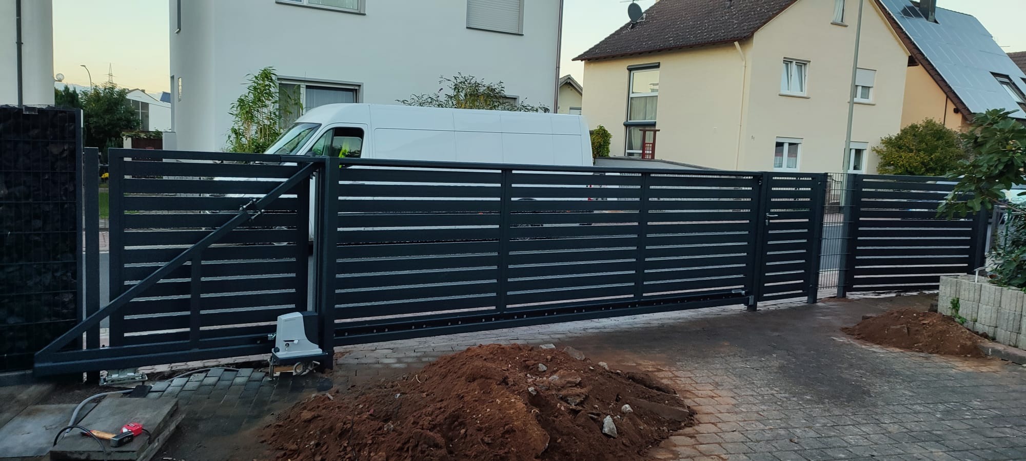 Nowoczesna, przesuwna brama wjazdowa z poziomych profili, automatyka bramy widoczna u dołu, w tle domy jednorodzinne i biały samochód dostawczy, świeżo usypana pryzma ziemi na kostce brukowej.
