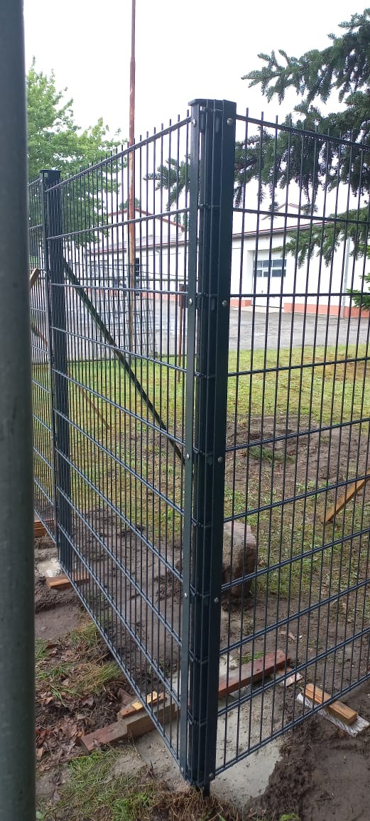 Fragment nowo zamontowanego, ciemnoszarego ogrodzenia panelowego z widocznymi drewnianymi podkładkami poziomującymi, na tle budynku z białej cegły i zieleni.