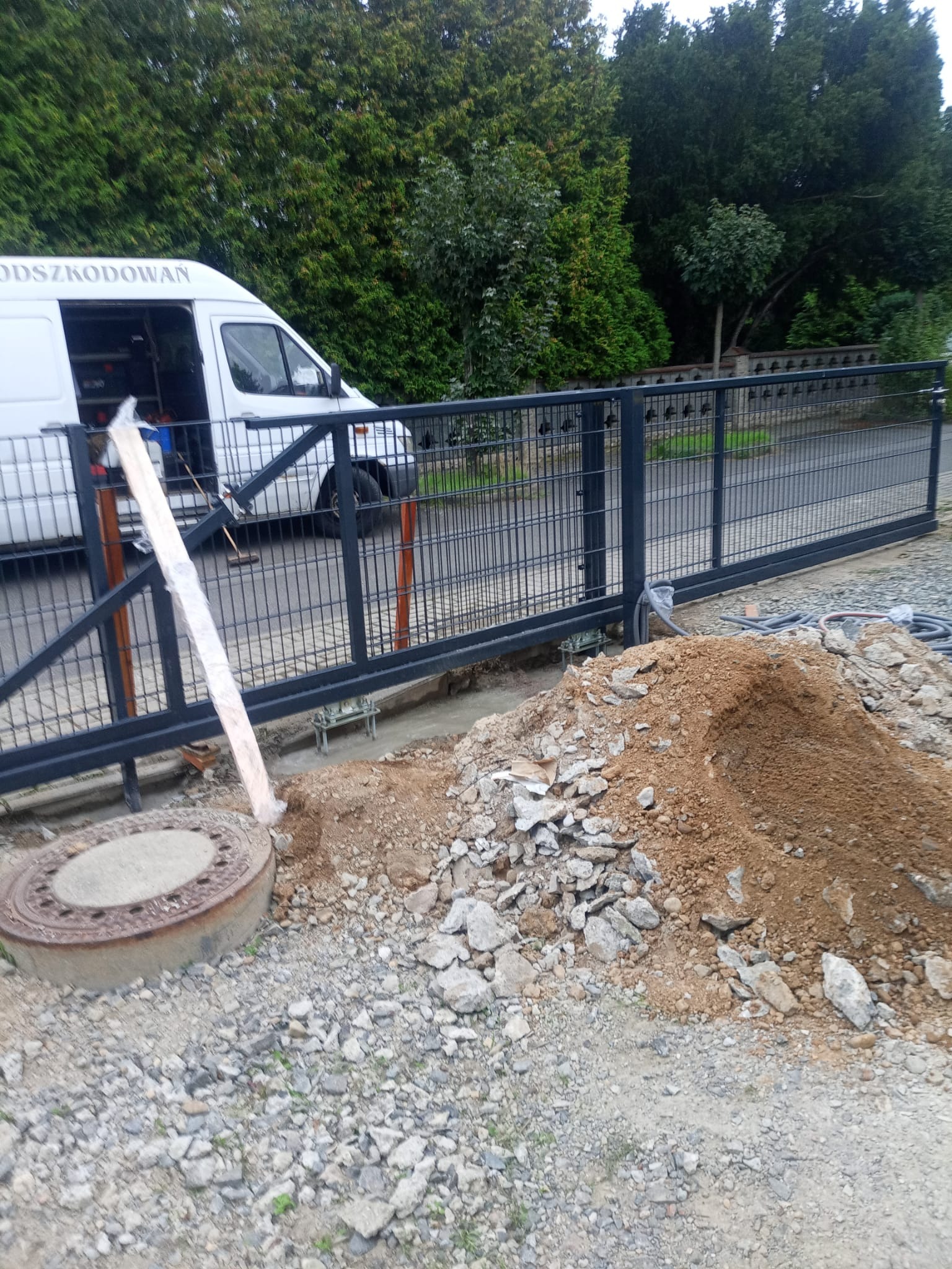 Montaż przesuwnej bramy z paneli metalowych w trakcie prac ziemnych, widoczny samochód dostawczy i sterta ziemi.