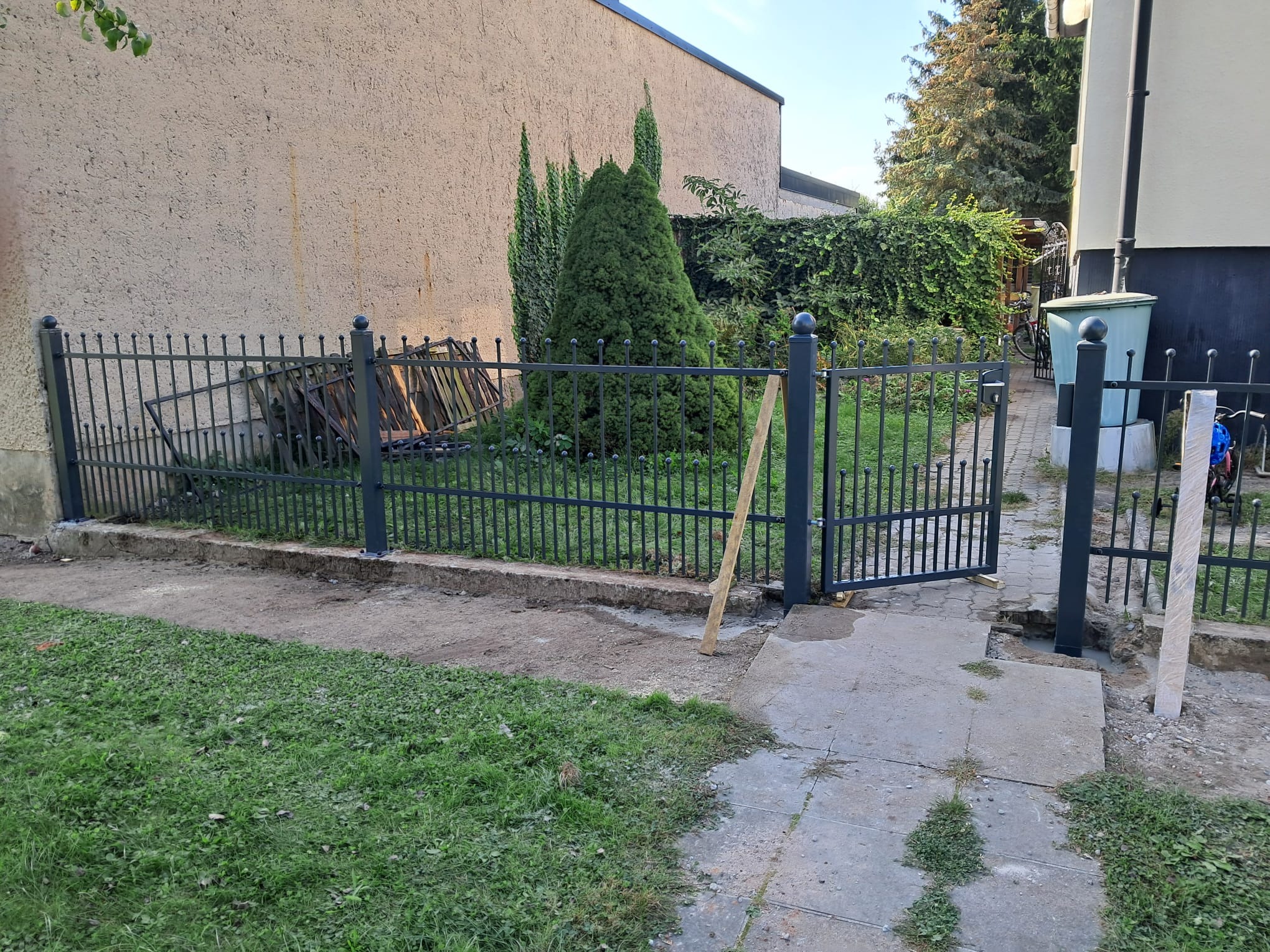 Montaż ogrodzenia metalowego z otwartą furtką na posesji z zielenią, widoczne ślady prac ziemnych i betonowania słupków.