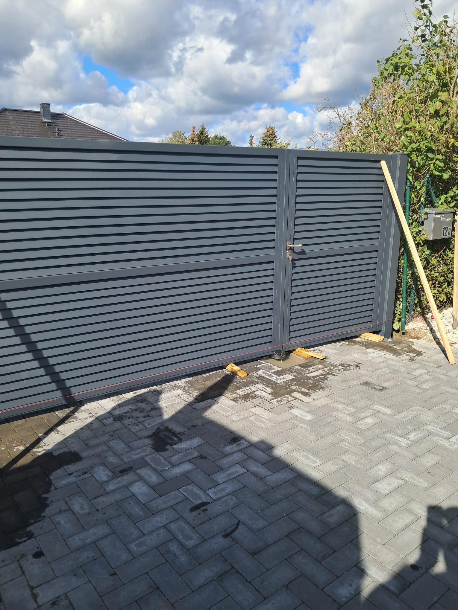 Szara brama wjazdowa z poziomych paneli, widoczne drewniane podpórki i poziomica, kostka brukowa na podjeździe, częściowo zachmurzone niebo.