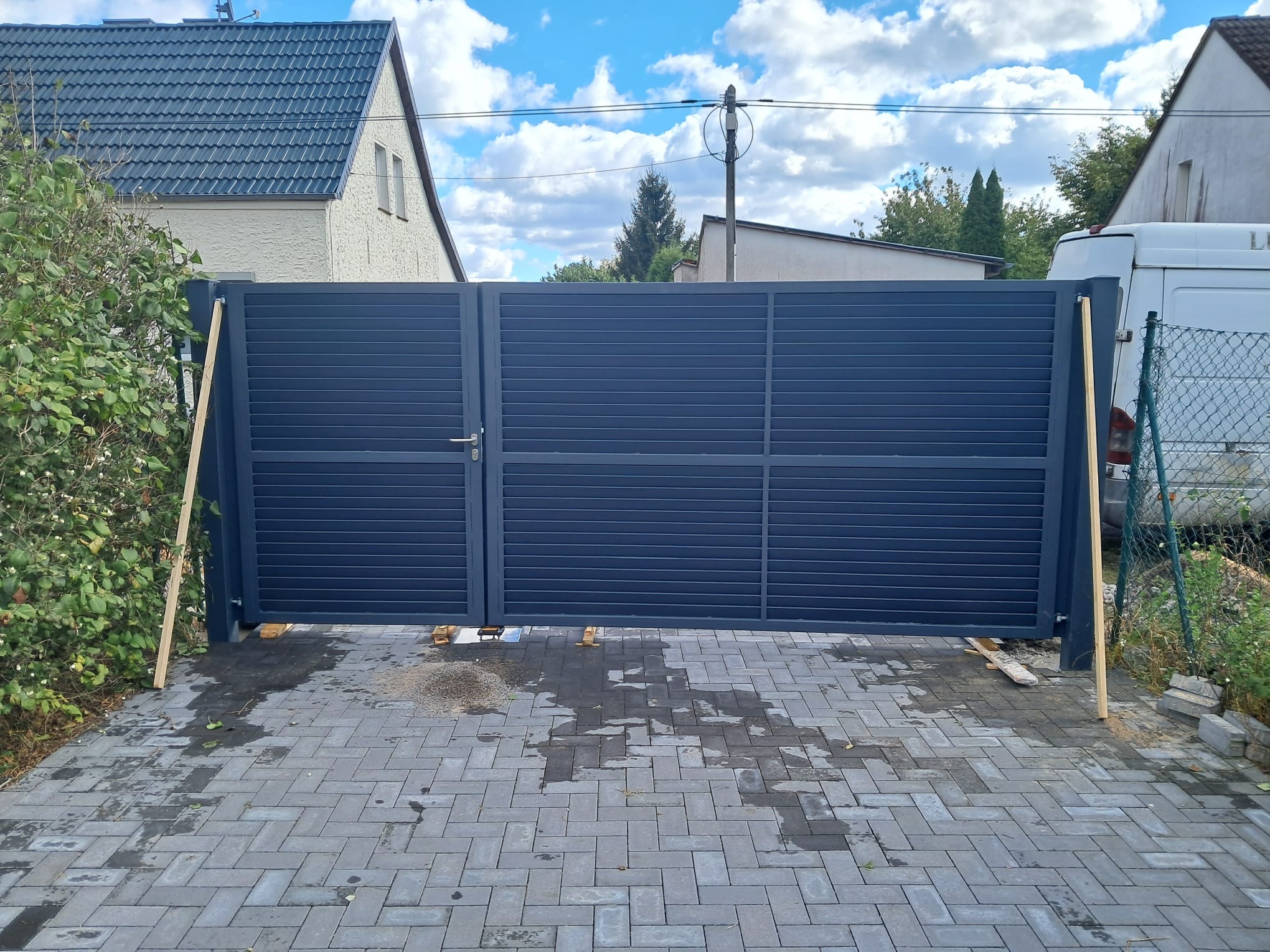 Nowoczesna, grafitowa brama przesuwna z poziomych paneli, częściowo zamontowana, podparta drewnianymi belkami, na tle kostki brukowej i budynku mieszkalnego.