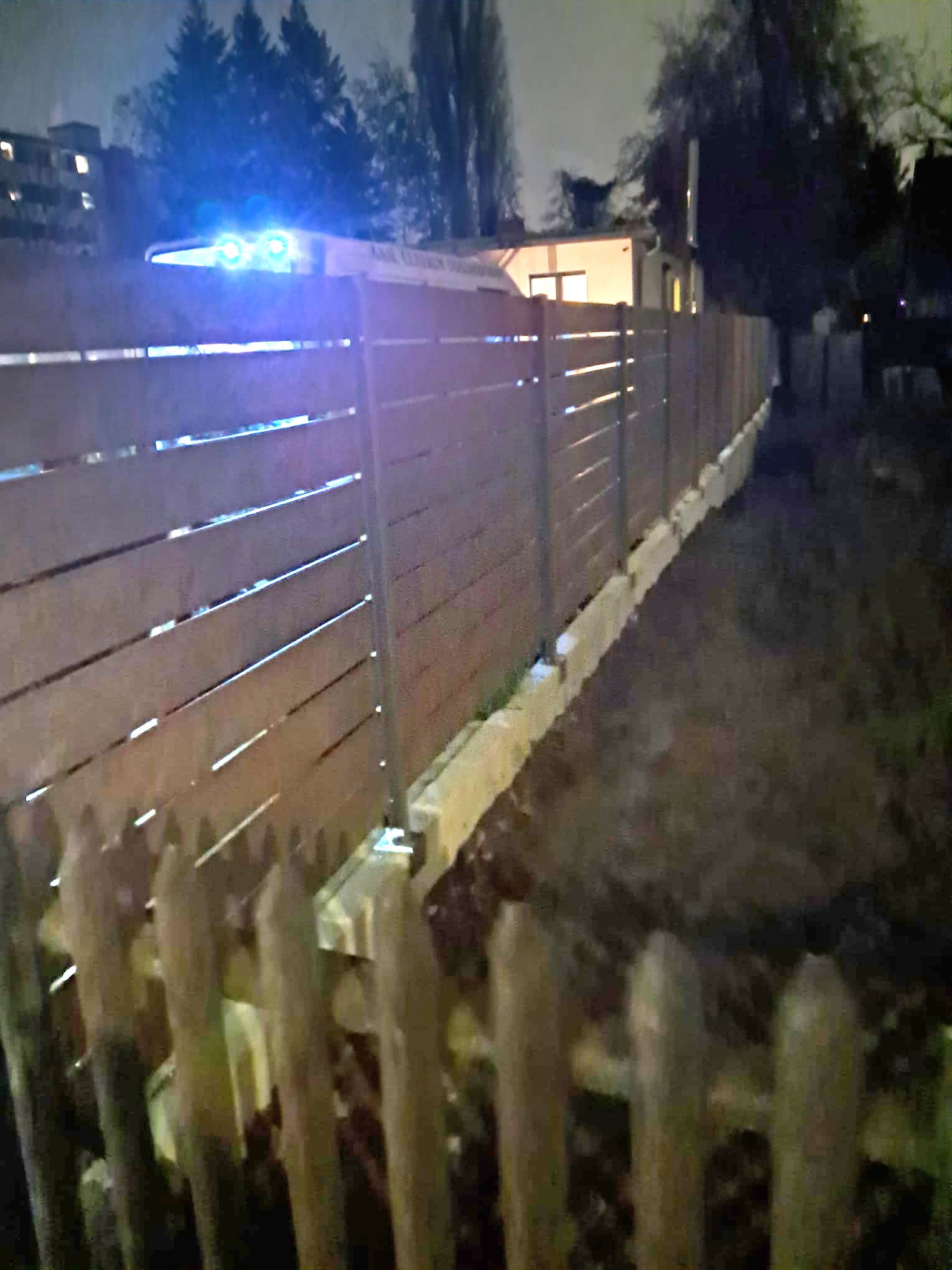 Nowoczesne ogrodzenie z poziomych desek i metalowych słupków, oświetlone niebieskim światłem w nocy, z widocznym fragmentem starego drewnianego płotu.