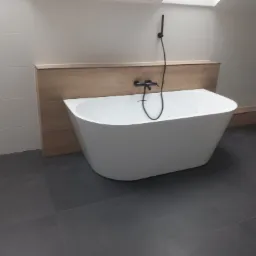 Nowoczesna, wolnostojąca wanna z czarną armaturą na tle jasnych płytek i drewnianej zabudowy w wyremontowanej łazience z szarą podłogą z płytek.