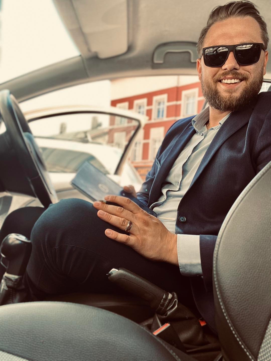 Uśmiechnięty mężczyzna w garniturze i okularach przeciwsłonecznych siedzi w samochodzie, trzyma tablet i patrzy w obiektyw.